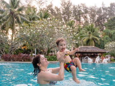 Beyaz bir kadın tatilde havuzda yüzerken oğlunu kucağına alıyor. Annesi küçük çocuğa yüzmeyi öğretiyor.