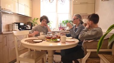 İki genç kardeş evde anneleriyle hamburger yiyorlar.