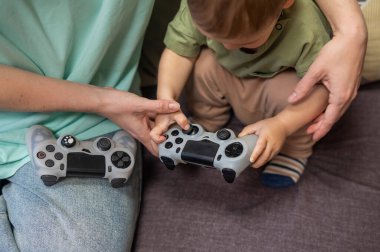 Beyaz kadın küçük oğluna konsol oynamayı öğretiyor. Oyun kolu ellerinde.