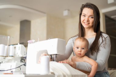 Genç bir kadın mutfakta dikiş dikiyor ve küçük bir çocuğu tutuyor. Annem küçük oğluna dikiş makinesi dikmeyi öğretiyor. Kendi kendini izole etme ödevi.