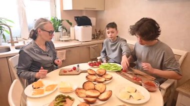 Orta yaşlı beyaz kadın iki oğluyla hamburger pişiriyor.