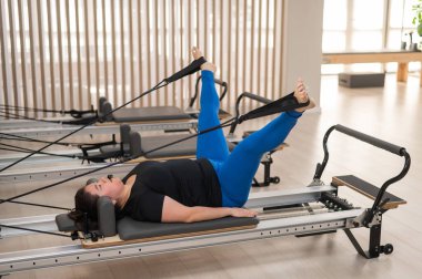 Aşırı kilolu beyaz kadın bir reformcu üzerinde pilates egzersizi yapıyor.