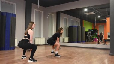 İki beyaz kadın dambıllarıyla çömeliyor.