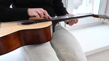 Genç adam pencere kenarında oturmuş akustik gitar çalıyor. Parmak Biçimi