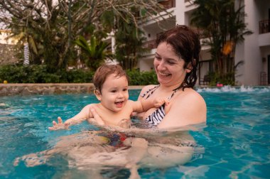 Beyaz bir kadın tatilde havuzda yüzerken oğlunu kucağına alıyor. Annesi küçük çocuğa yüzmeyi öğretiyor.