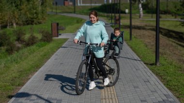 Beyaz kadın bisiklete biniyor. Yanında da oğlu var. Parkta çocuk koltuğunda oturuyor.