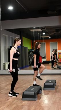 İki beyaz kadın dambıllarıyla adım egzersizi yapıyor.