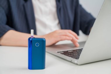İş kadını dizüstü bilgisayarla uğraşırken elektronik sigara içiyor. Ön planda tütün ısıtma sistemi