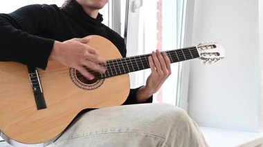 Genç adam pencere kenarında oturmuş akustik gitar çalıyor. Parmak Biçimi