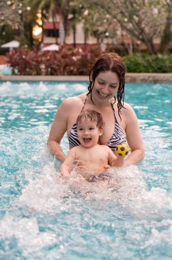 Beyaz bir kadın tatilde havuzda yüzerken oğlunu kucağına alıyor. Annesi küçük çocuğa yüzmeyi öğretiyor.