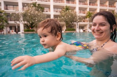 Beyaz bir kadın tatilde havuzda yüzerken oğlunu kucağına alıyor. Annesi küçük çocuğa yüzmeyi öğretiyor.