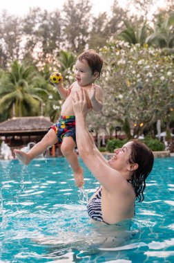 Beyaz bir kadın tatilde havuzda yüzerken oğlunu kucağına alıyor. Annesi küçük çocuğa yüzmeyi öğretiyor.
