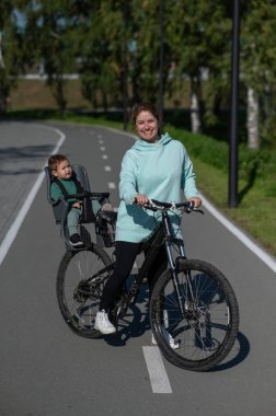 Beyaz bir kadın, parkta çocuk koltuğunda arkasında oturan bebekle bisiklete biniyor. Dikey fotoğraf