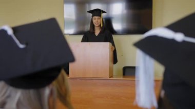 Genç bir kadın kürsüde duruyor ve sınıf arkadaşlarına mezuniyet konuşması yapıyor.
