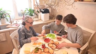 Orta yaşlı beyaz kadın iki oğluyla hamburger pişiriyor.