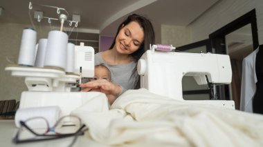 Genç bir kadın mutfakta dikiş dikiyor ve küçük bir çocuğu tutuyor. Annem küçük oğluna dikiş makinesi dikmeyi öğretiyor. Kendi kendini izole etme ödevi.