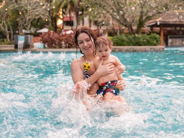 Beyaz bir kadın tatilde havuzda yüzerken oğlunu kucağına alıyor. Annesi küçük çocuğa yüzmeyi öğretiyor.