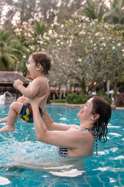 Beyaz bir kadın tatilde havuzda yüzerken oğlunu kucağına alıyor. Annesi küçük çocuğa yüzmeyi öğretiyor.
