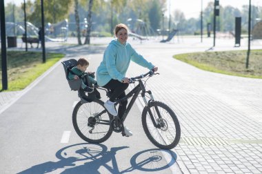 Beyaz kadın bisiklete biniyor. Yanında da oğlu var. Parkta çocuk koltuğunda oturuyor.