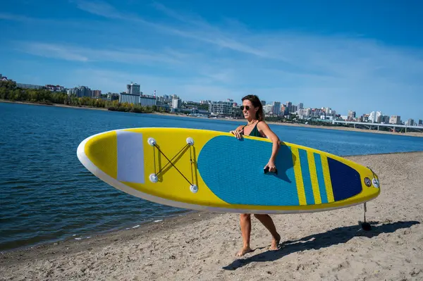 Beyaz bir kadın sahilde yürüyor ve şehirdeki nehirde bir yiyecek panosu taşıyor. Yaz sporu