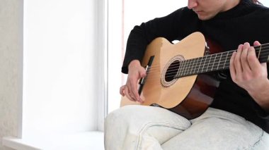 Genç adam pencere kenarında oturmuş akustik gitar çalıyor. Parmak Biçimi