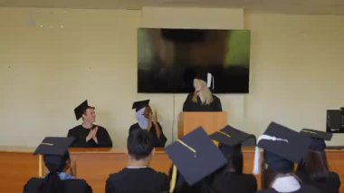 Genç bir kadın kürsüde duruyor ve sınıf arkadaşlarına mezuniyet konuşması yapıyor.