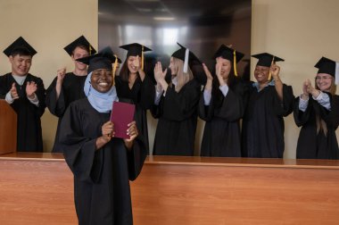 Tesettürlü mutlu Afrikalı kadın diploma alıyor ve sınıf arkadaşları onu alkışlıyor.