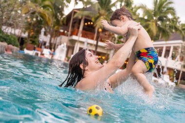 Beyaz bir kadın tatilde havuzda yüzerken oğlunu kucağına alıyor. Annesi küçük çocuğa yüzmeyi öğretiyor.