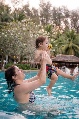 Beyaz bir kadın tatilde havuzda yüzerken oğlunu kucağına alıyor. Annesi küçük çocuğa yüzmeyi öğretiyor.