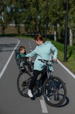 Beyaz bir kadın, parkta çocuk koltuğunda arkasında oturan bebekle bisiklete biniyor. Dikey fotoğraf
