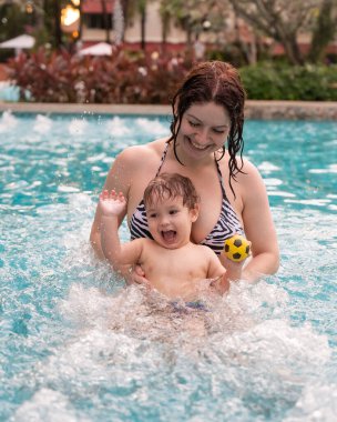 Beyaz bir kadın tatilde havuzda yüzerken oğlunu kucağına alıyor. Annesi küçük çocuğa yüzmeyi öğretiyor.