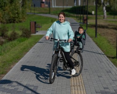 Beyaz kadın bisiklete biniyor. Yanında da oğlu var. Parkta çocuk koltuğunda oturuyor.
