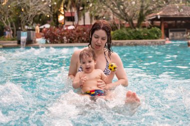 Beyaz bir kadın tatilde havuzda yüzerken oğlunu kucağına alıyor. Annesi küçük çocuğa yüzmeyi öğretiyor.