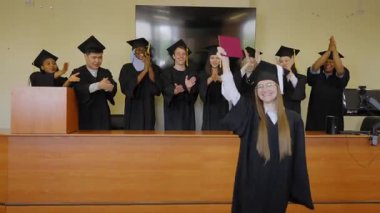 Sınıf arkadaşları onu alkışlarken, elinde diploma tutan beyaz bir kadın.