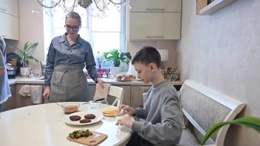 Orta yaşlı beyaz kadın iki oğluyla hamburger pişiriyor.