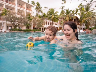 Beyaz bir kadın tatilde havuzda yüzerken oğlunu kucağına alıyor. Annesi küçük çocuğa yüzmeyi öğretiyor.