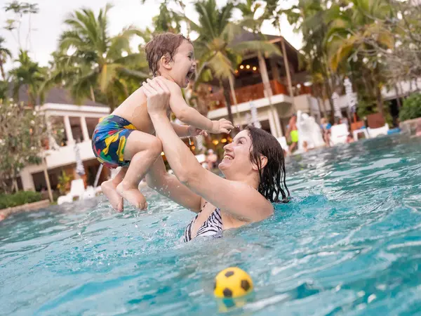 Beyaz bir kadın tatilde havuzda yüzerken oğlunu kucağına alıyor. Annesi küçük çocuğa yüzmeyi öğretiyor.