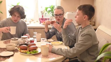 Orta yaşlı beyaz bir kadın iki oğluyla yemek yiyor.