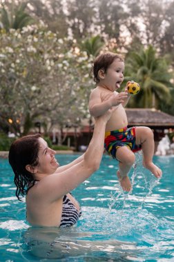 Beyaz bir kadın tatilde havuzda yüzerken oğlunu kucağına alıyor. Annesi küçük çocuğa yüzmeyi öğretiyor.