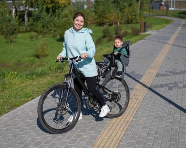 Beyaz kadın bisiklete biniyor. Yanında da oğlu var. Parkta çocuk koltuğunda oturuyor.
