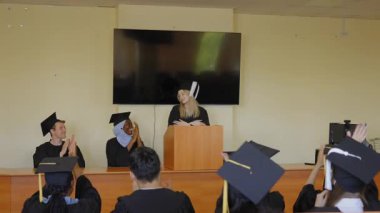 Genç bir kadın kürsüde duruyor ve sınıf arkadaşlarına mezuniyet konuşması yapıyor.