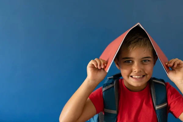 青い背景 コピースペースの上に立って頭の上に本を持つ遊び心のある白人少年の肖像画 変更されていない ライフスタイル 子供時代 学校の少年 幸せなコンセプト — ストック写真