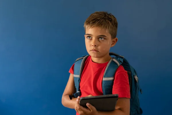 白种人男孩站在蓝色的背景上 带着数码平板电脑和书包 望着远方 复制空间 生活方式 技术和沉思的概念 — 图库照片