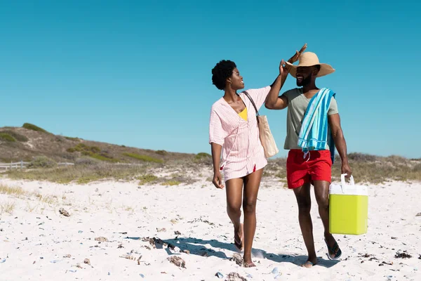 快乐的非洲裔美国女人在清澈的天空下走在海滩上 触摸着丈夫戴着的帽子 不变的 聚会的 度假的 自然的 毛巾的 凉爽的 享受的和夏天的概念 — 图库照片