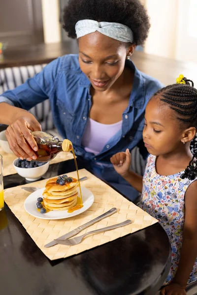 アフリカ系アメリカ人の女性が家で娘と朝食をしながらパンケーキに蜂蜜を注ぐ 変更されていない 一緒に 子供時代 食べ物 新鮮な ダイニングテーブルと家庭の概念 — ストック写真