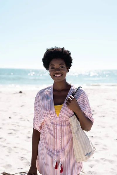 澄んだ空の下でビーチでバッグを運ぶアフロヘアでアフリカ系アメリカ人の中人女性を笑顔 コピースペース 変更されていない ライフスタイル 楽しみ 肖像画と夏のコンセプト — ストック写真
