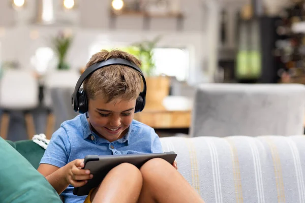 坐在沙发上 戴着耳机和数字平板电脑的白人男孩笑着 不变的生活方式 技术和快乐的概念 — 图库照片