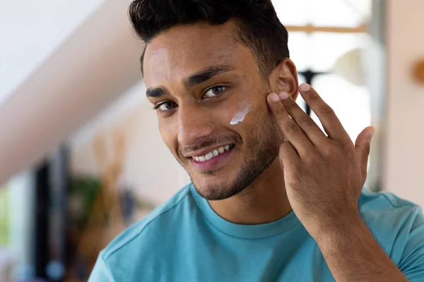 Hombre Birracial Sonriente Mirándose Espejo Del Baño Aplicando Crema Facial —  Fotos de Stock