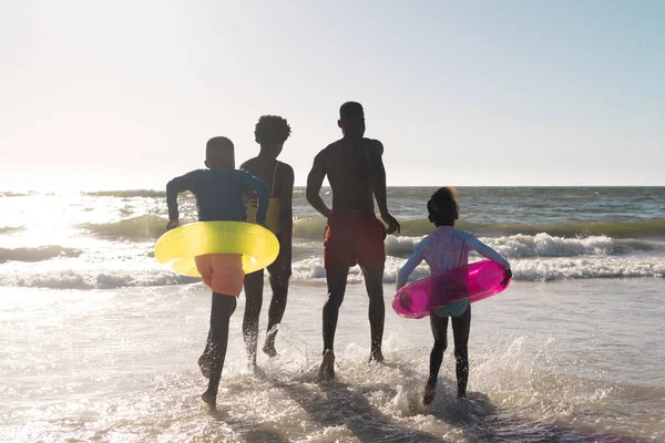 澄んだ空を背景に海を走る明るいアフリカ系アメリカ人の両親と子供たちの後ろ姿 コピースペース 変更されていない 一緒に 子供時代 水泳フロート 楽しみ — ストック写真