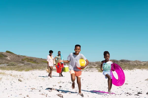 アフリカ系アメリカ人の遊び心のある子供たちが走り 両親は澄んだ青空の下で砂浜を歩いています コピースペース 変更されていない 一緒に ピクニック ボール 水泳フロート 楽しみ — ストック写真